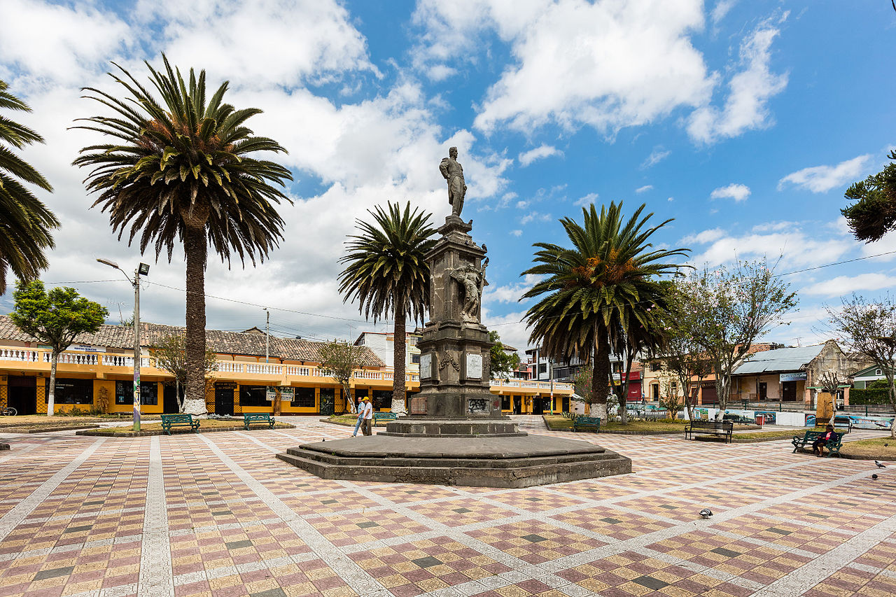 Das Bild fokussiert die Statue auf dem Plaza de Armas in Ibarra, Ecuador. Auf dem Platz verteilt stehen Palmen, außerhalb stehen einige farbenfrohe Gebäude. Der Platz hat einen kompletten Mosaikboden.